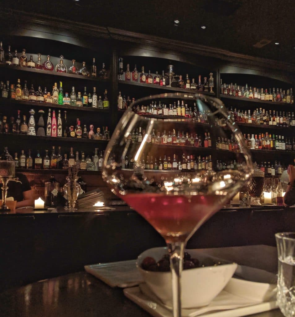 Picture of cocktail against bar with hundreds of bottles behind it. 